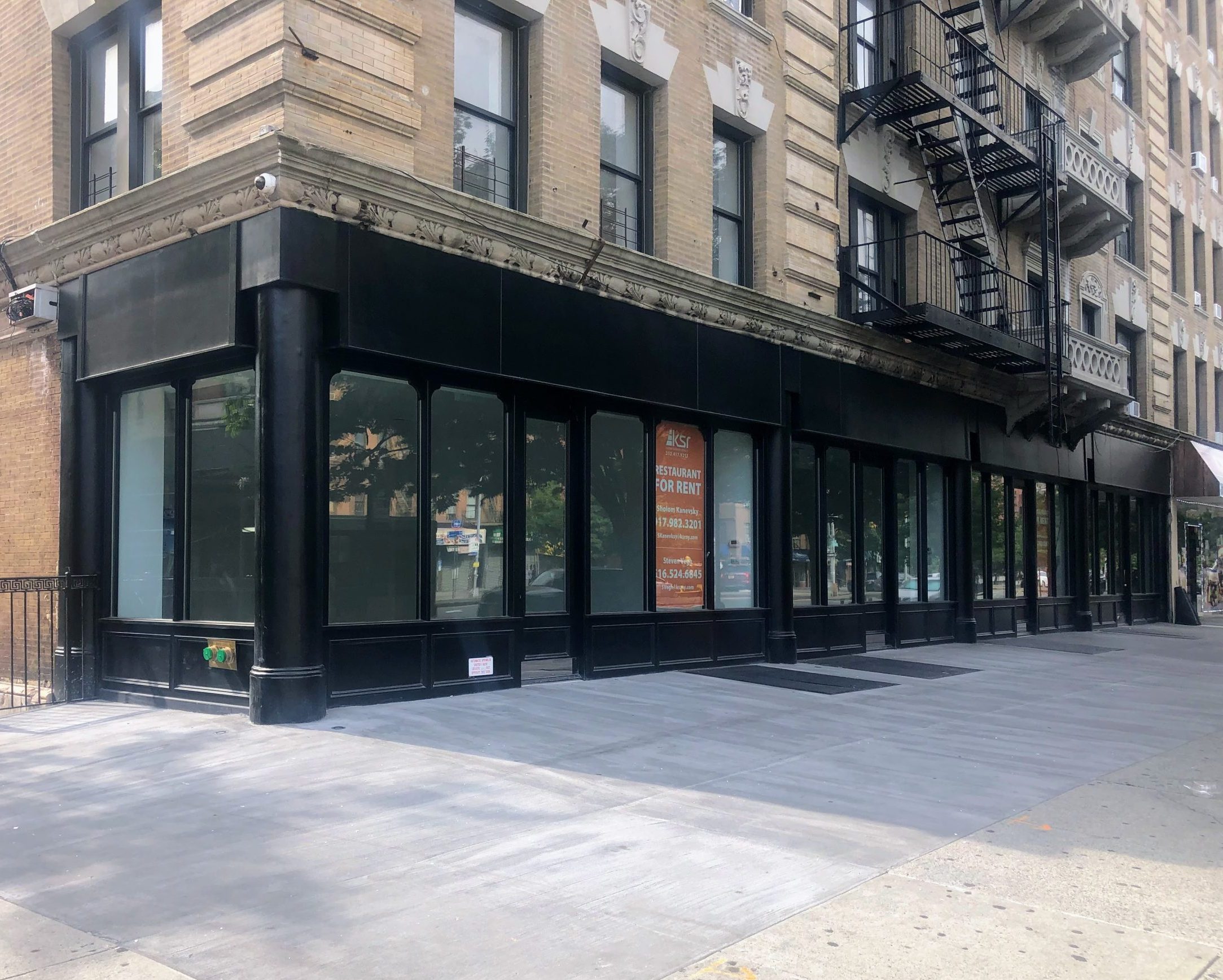 Exterior view of a storefront with large glass windows and a black framing on a city street, featuring a 'For Rent' sign in the window.